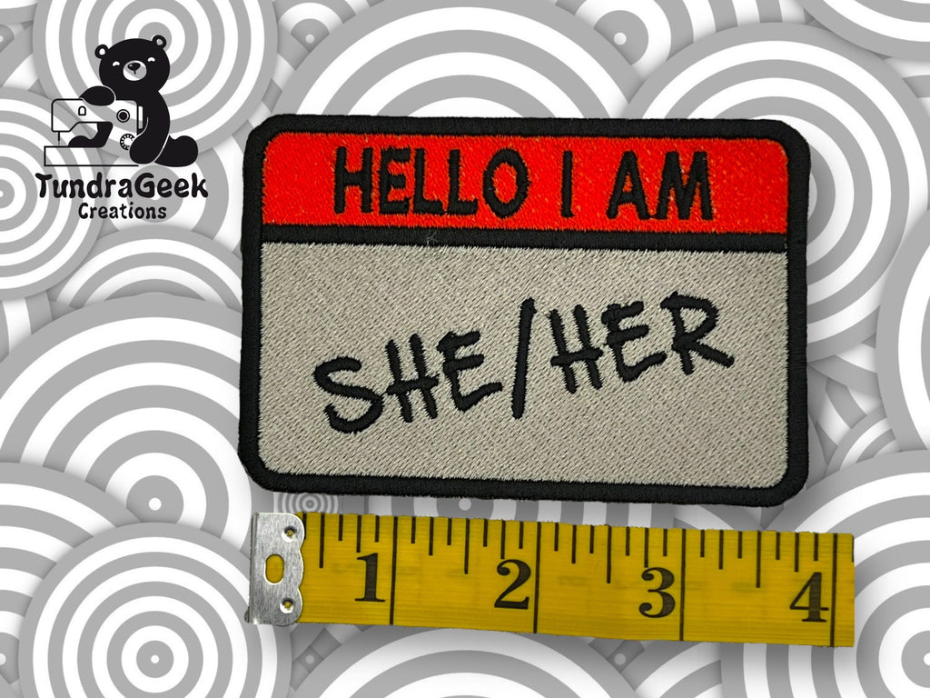 A rectangular embroidered patch reads HELLO I AM SHE/HER above a yellow measuring tape showing about 4 inches, with a TundraGeek Creations logo in the top left corner.