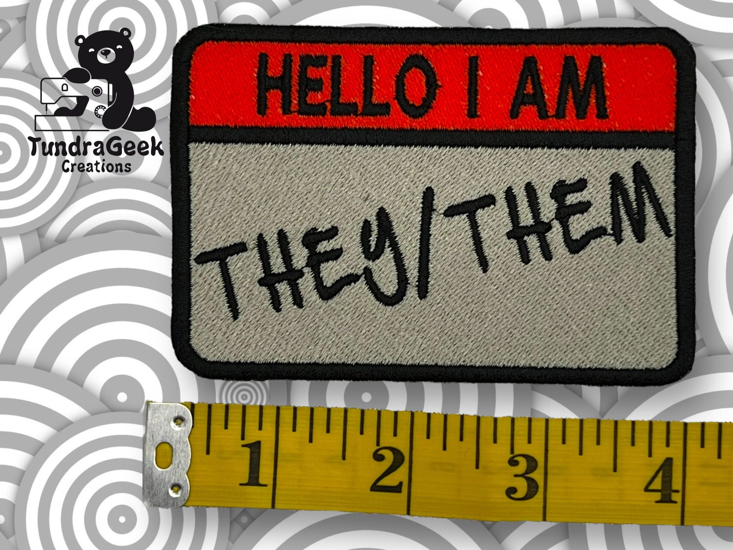 A fabric patch reads HELLO I AM THEY/THEM with a measuring tape showing its size, and a TundraGeek Creations logo in the top left corner.