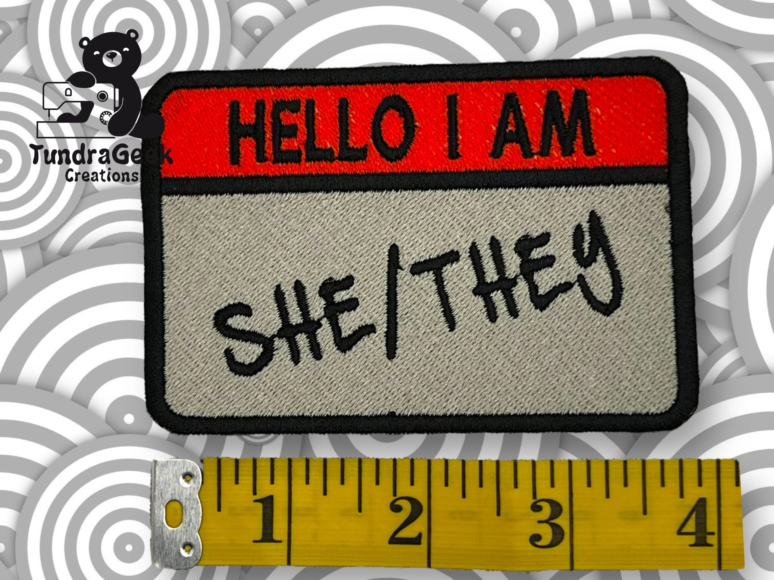 A fabric patch resembling a Hello I Am name tag with She/They written on it, next to a yellow measuring tape and a TundraGeek Creations logo in the background.