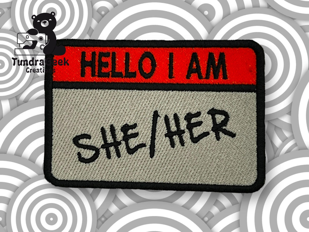 An embroidered patch designed like a Hello I Am name tag with SHE/HER written in black letters, set against a background with gray circular patterns.