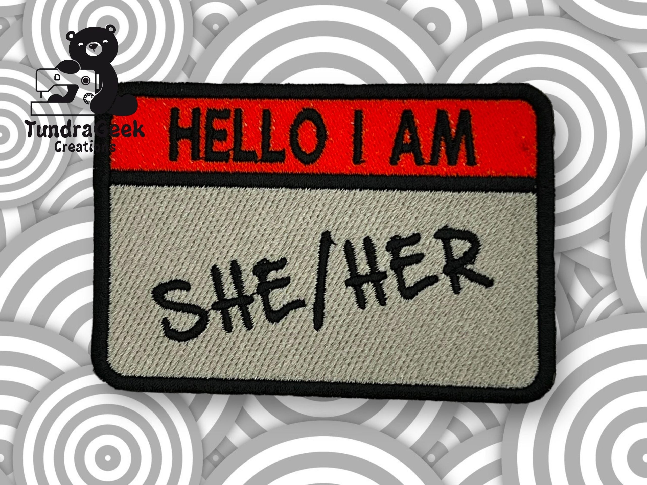 An embroidered patch designed like a Hello I Am name tag with SHE/HER written in black letters, set against a background with gray circular patterns.
