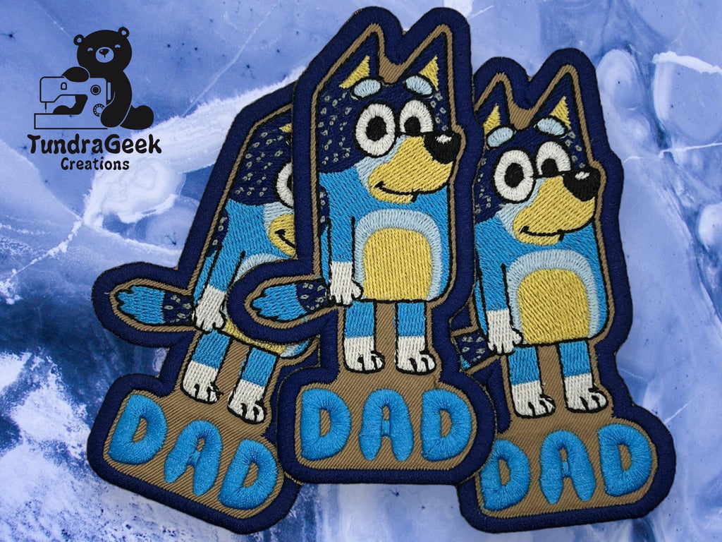 Three embroidered patches of a cartoon blue dog labeled DAD are displayed on a blue background, with a TundraGeek Creations logo in the top left corner.
