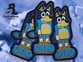 Three embroidered patches of a cartoon blue dog labeled DAD are displayed on a blue background, with a TundraGeek Creations logo in the top left corner.