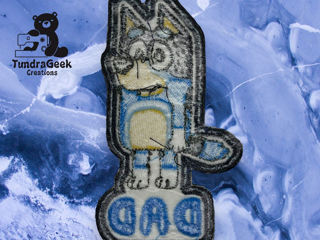 Embroidered patch featuring a cartoon dog and the word DAD on a blue marbled background, with a TundraGeek Creations logo in the top left corner.