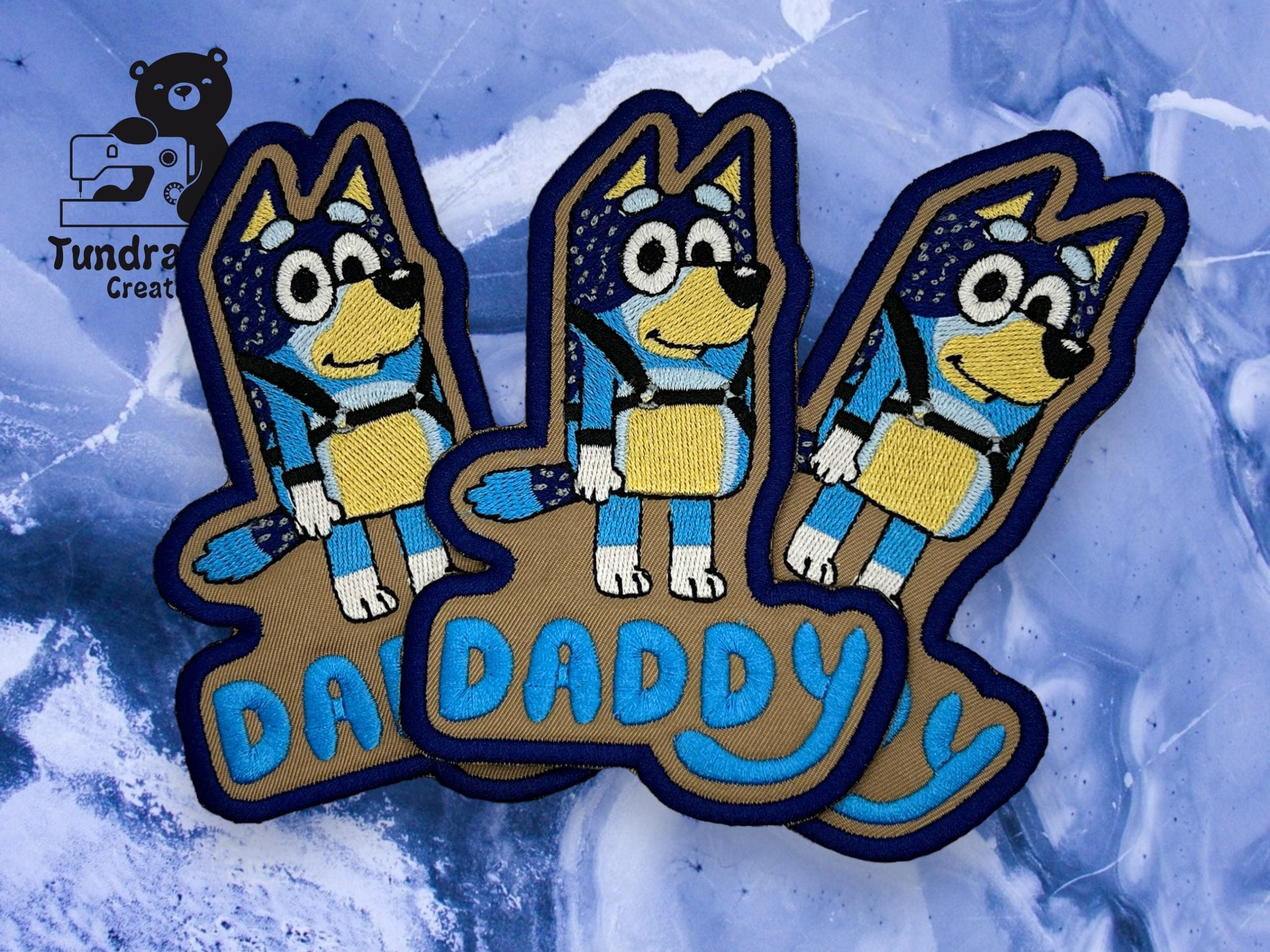Three embroidered patches feature a cartoon blue dog with the word DADDY in blue text, placed on a blue and white background. Tundra Creative logo is in the top left corner.