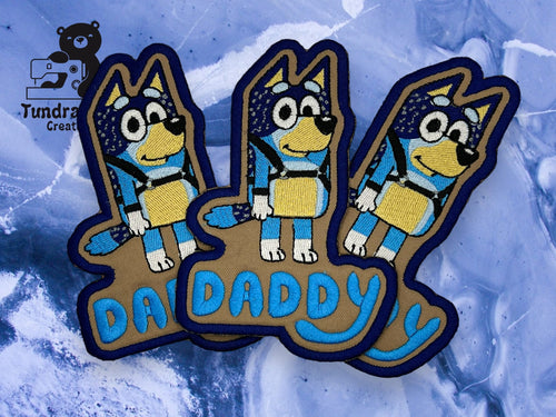 Three embroidered patches feature a cartoon blue dog with the word DADDY in blue text, placed on a blue and white background. Tundra Creative logo is in the top left corner.