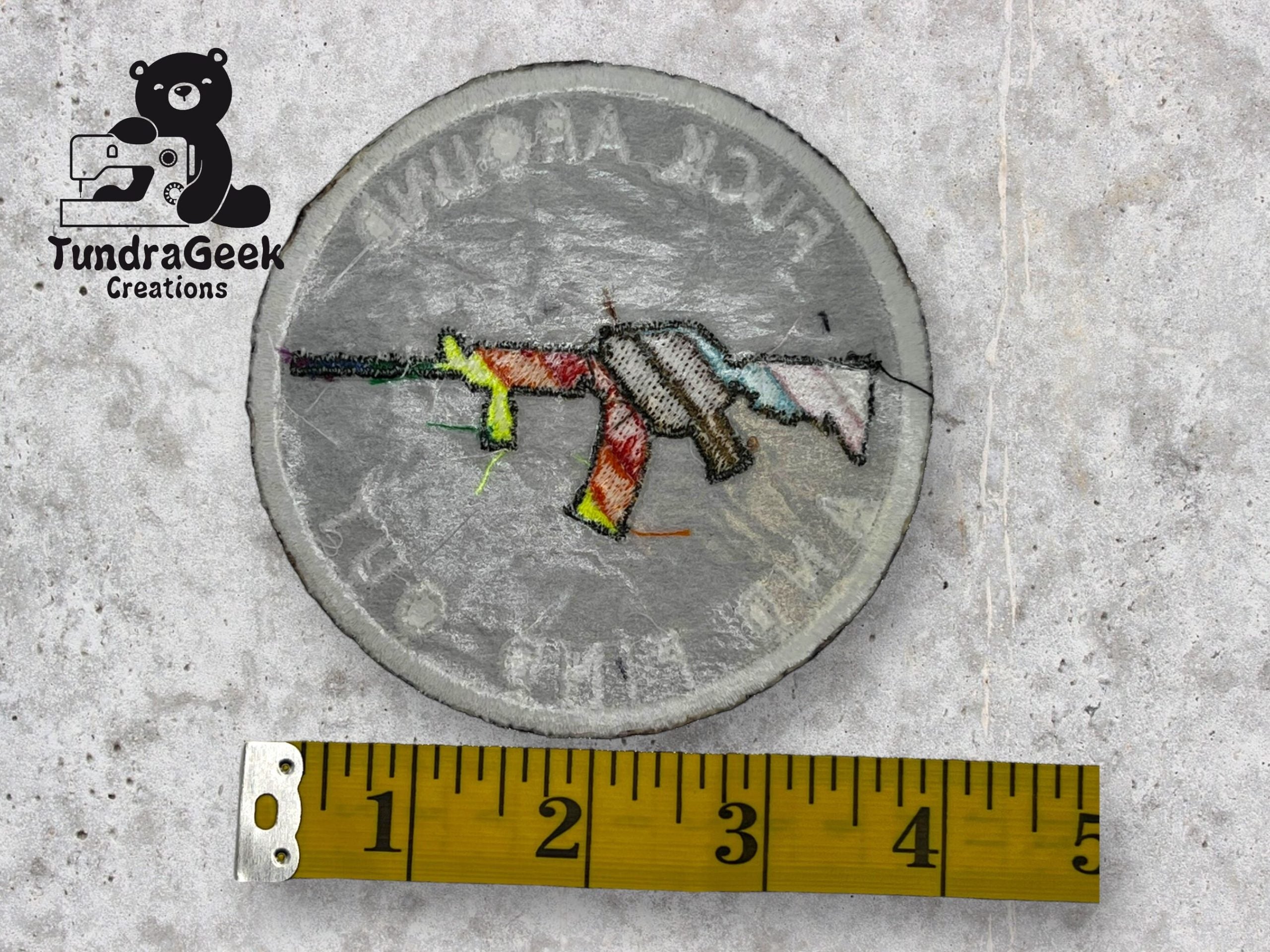 A handmade circular patch with a colorful rifle design and text around the edge is shown next to a yellow measuring tape on a concrete surface.