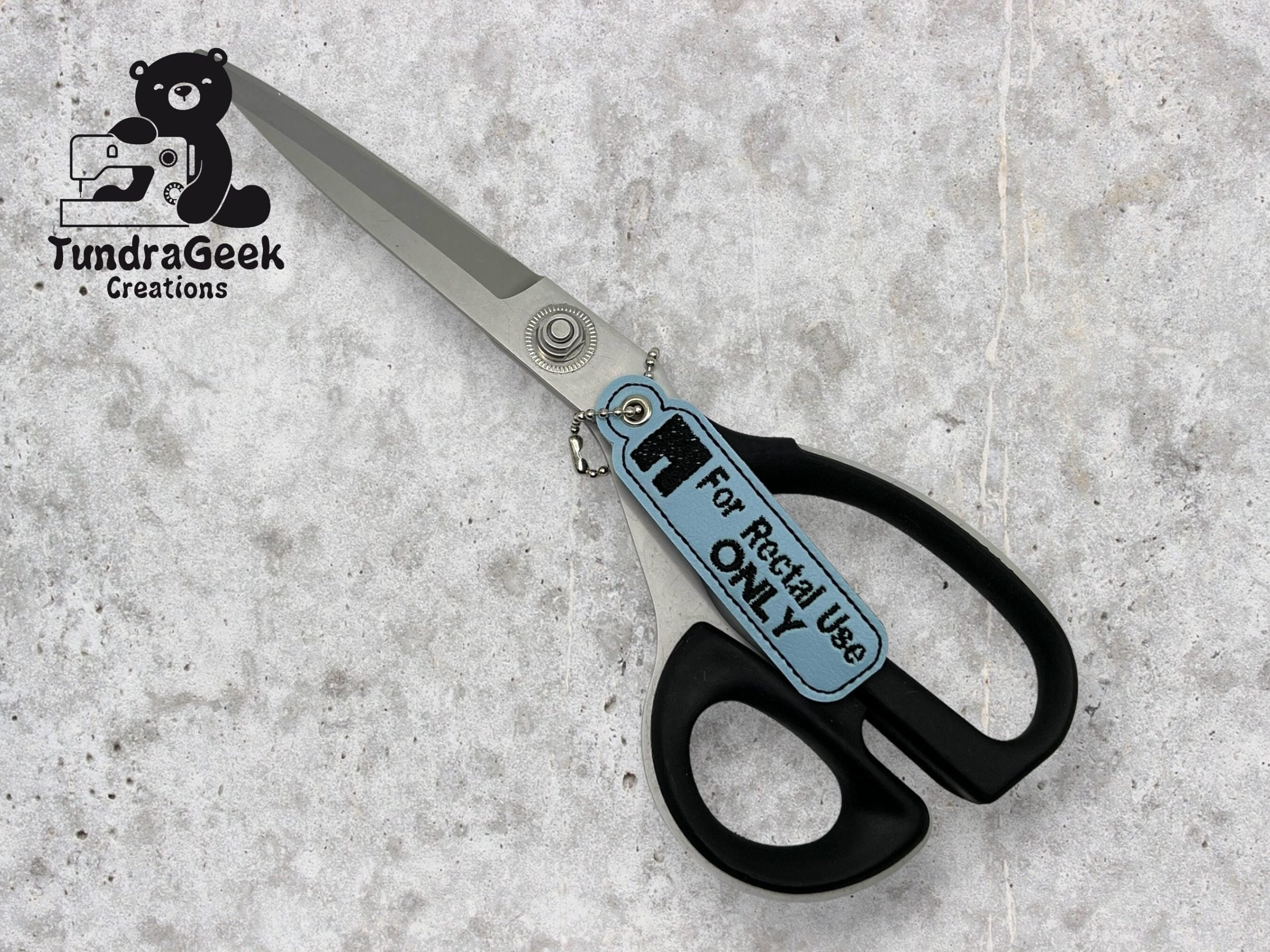 A pair of black-handled scissors with a tag reading For Rectal Use ONLY lies on a gray surface. Logo of TundraGeek Creations is visible in the top left corner.
