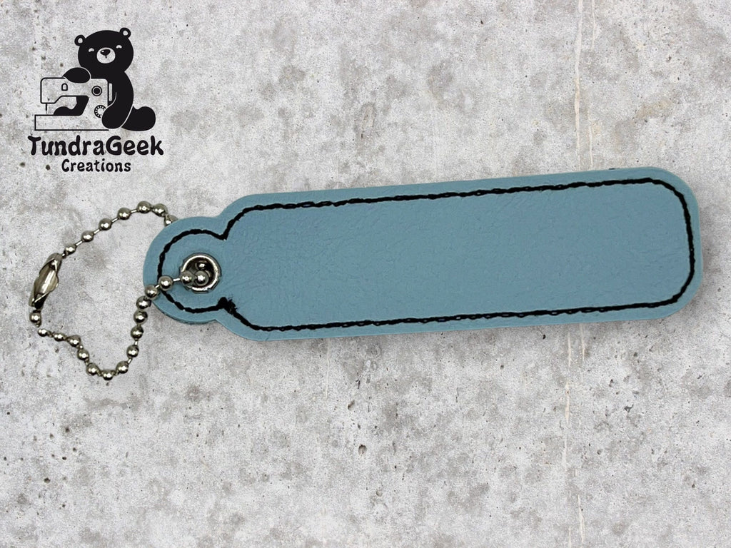 A blue leather keychain with black stitching and a metal ball chain, lying on a light textured surface. TundraGeek Creations logo is in the top left corner.