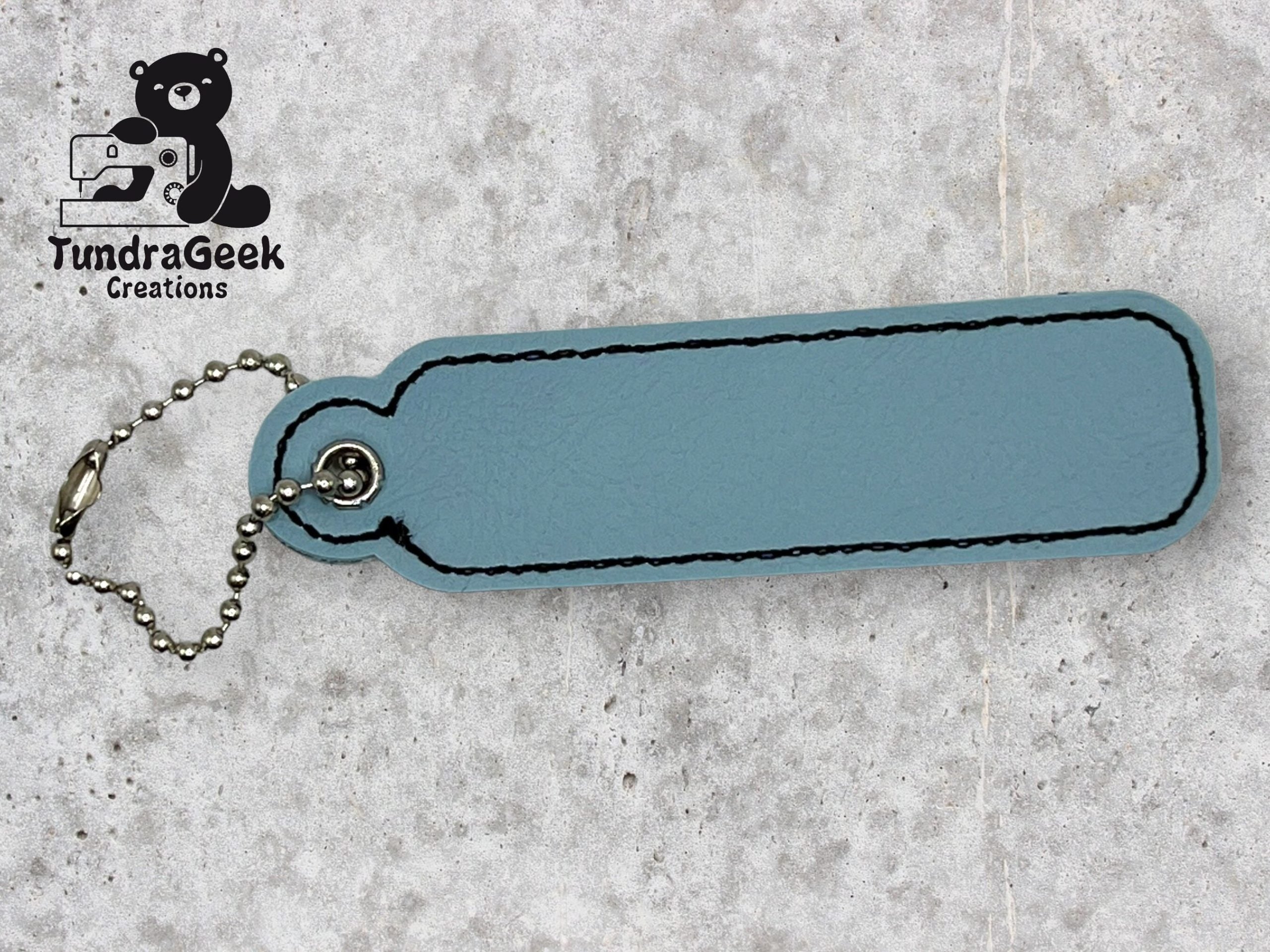 A blue leather keychain with black stitching and a metal ball chain, lying on a light textured surface. TundraGeek Creations logo is in the top left corner.