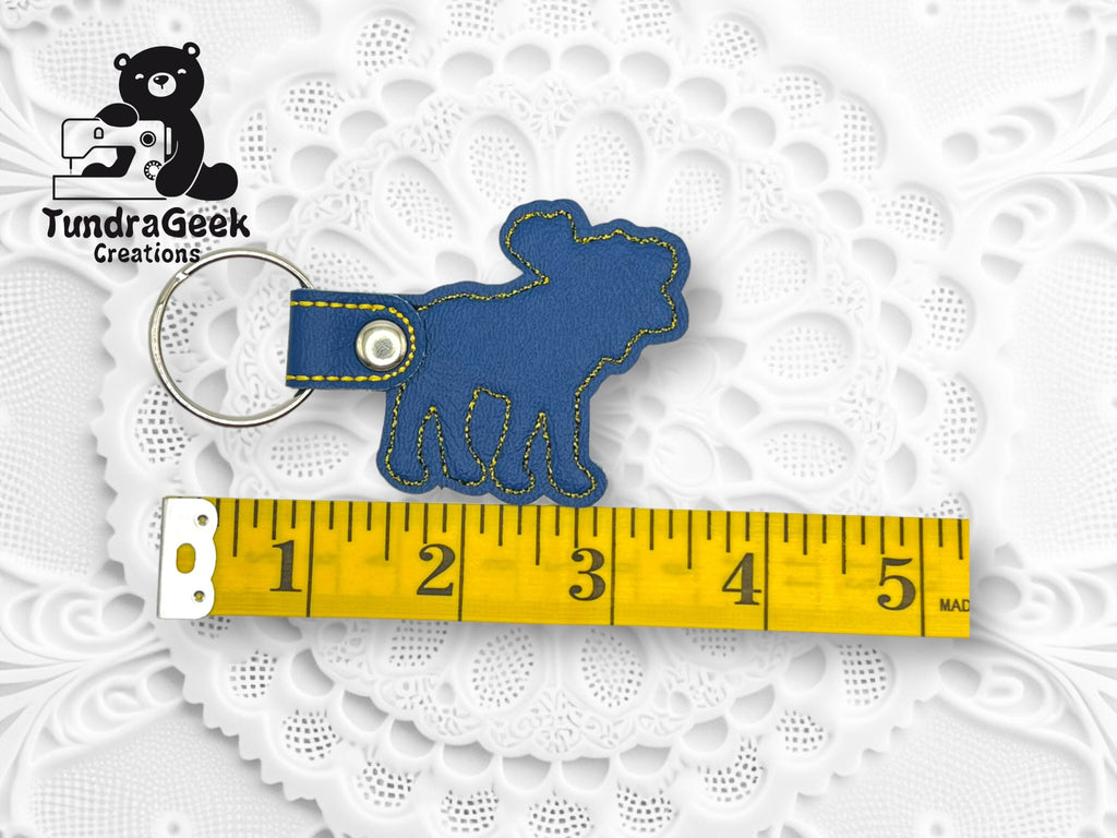 Blue bear-shaped keychain with yellow stitching, placed next to a yellow measuring tape showing a length of about 3 inches, on a white lace-patterned background.