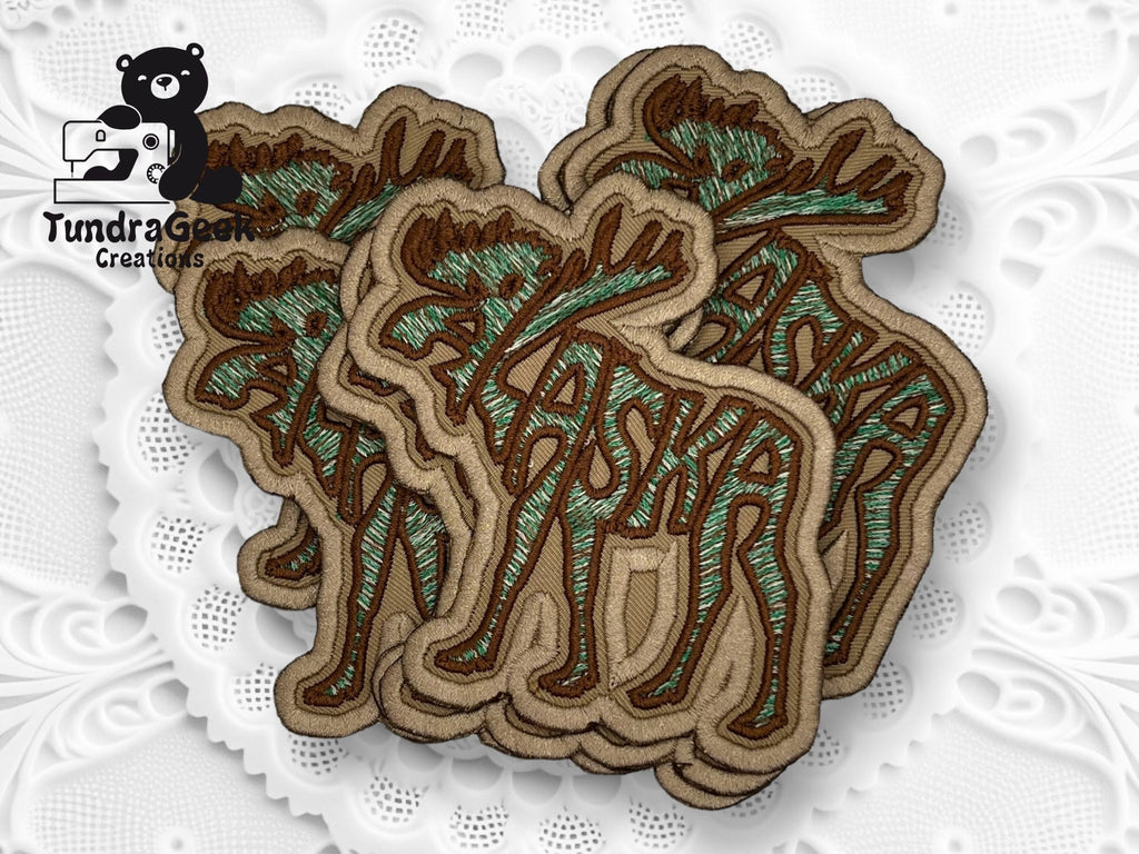 A stack of embroidered patches spelling Alaska in brown and green thread with trees incorporated into the design, displayed on a white textured background.