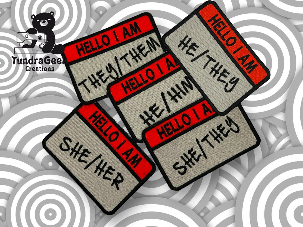 Five embroidered name tag patches display different pronouns: they/them, he/they, he/him, she/her, and she/they. The TundraGee Creations logo is in the top left corner.