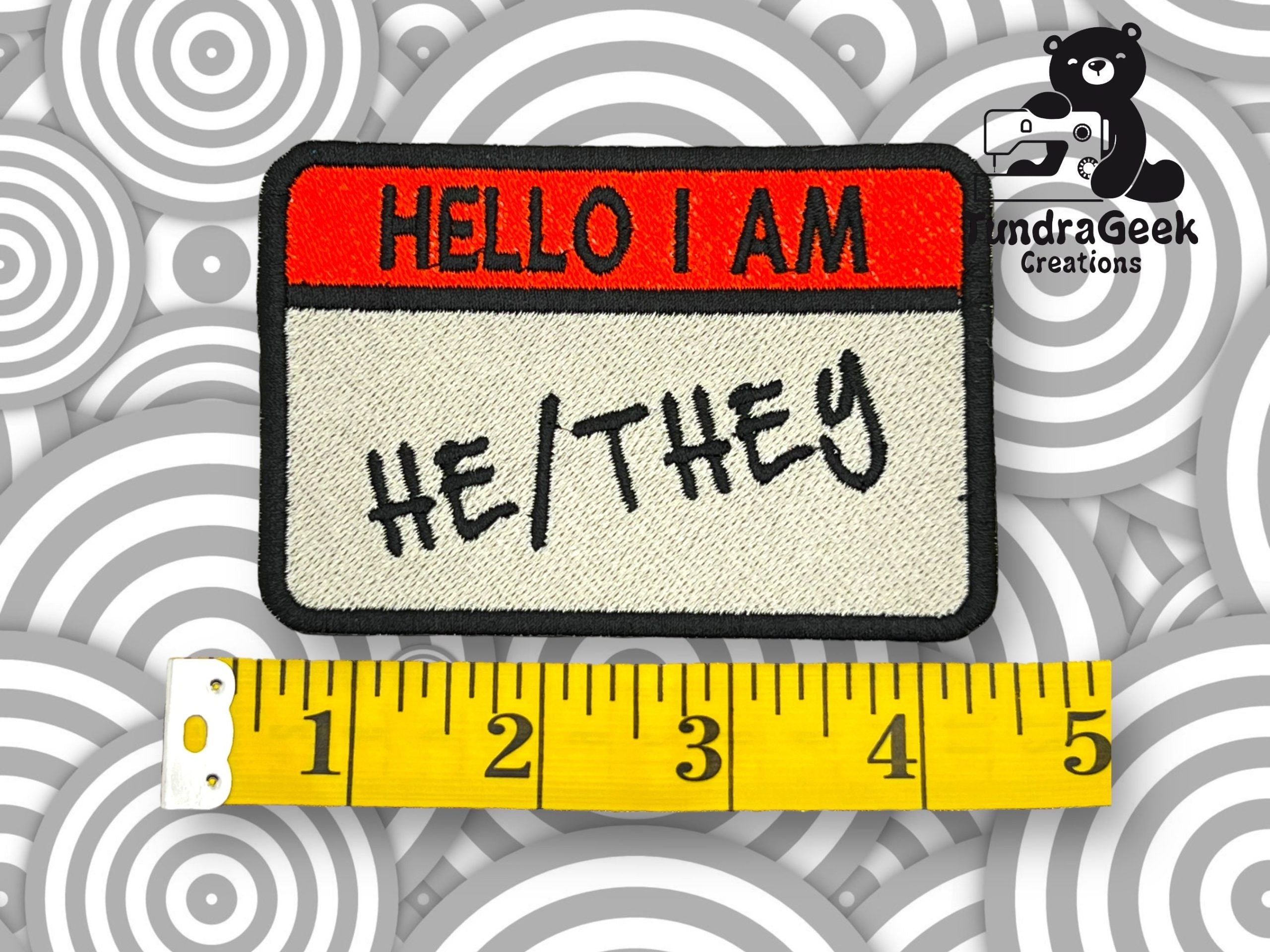 A fabric patch reads HELLO I AM HE/THEY beside a yellow measuring tape showing about 4 inches, with a bear and SandraGeek Creations logo in the top right corner.