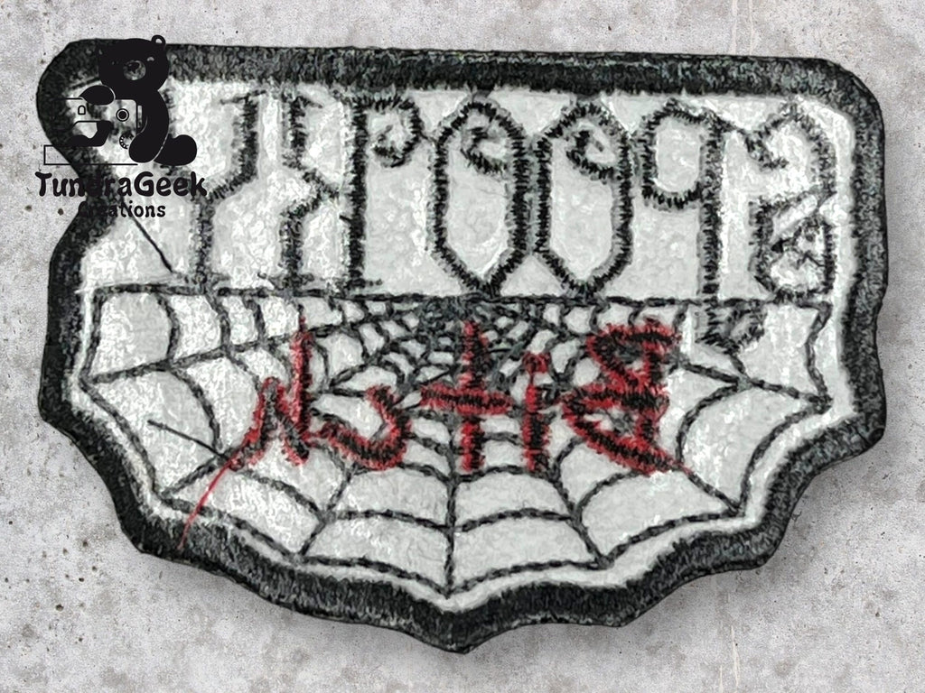 A patch shaped like a spider web with text in black and red embroidery on a white background, and a small black logo with a bear and TunaGeek Creations in the corner.