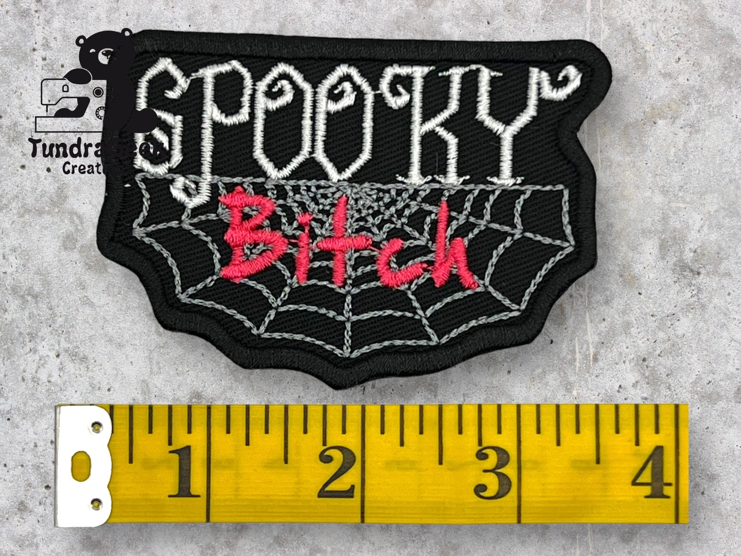 An embroidered patch reading Spooky Bitch with a spiderweb design, placed above a yellow measuring tape showing a length of about 3 inches.