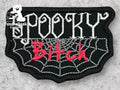 A black patch with a spiderweb design displays the words Spooky Bitch in white and pink embroidered letters.