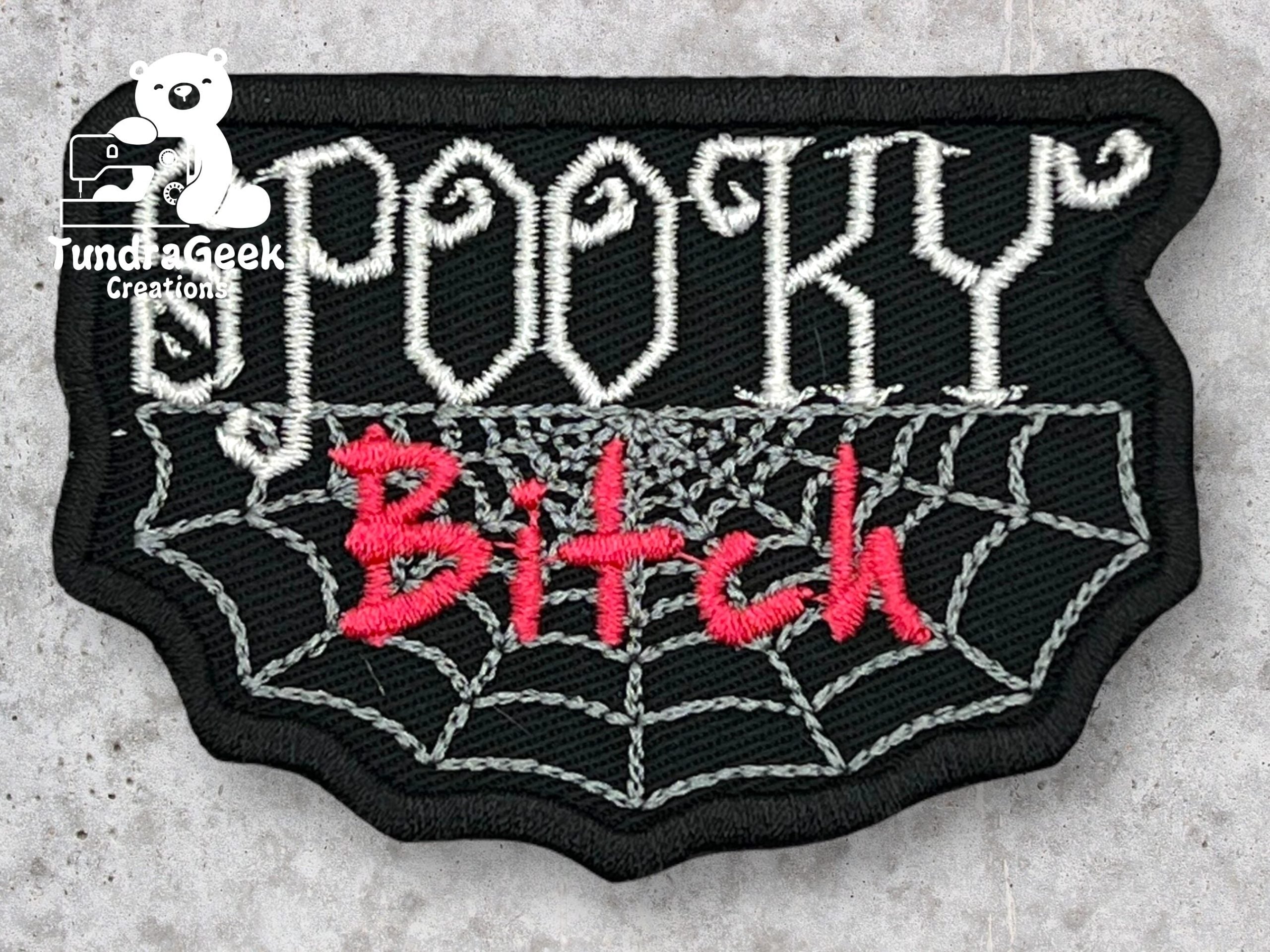 A black patch with a spiderweb design displays the words Spooky Bitch in white and pink embroidered letters.