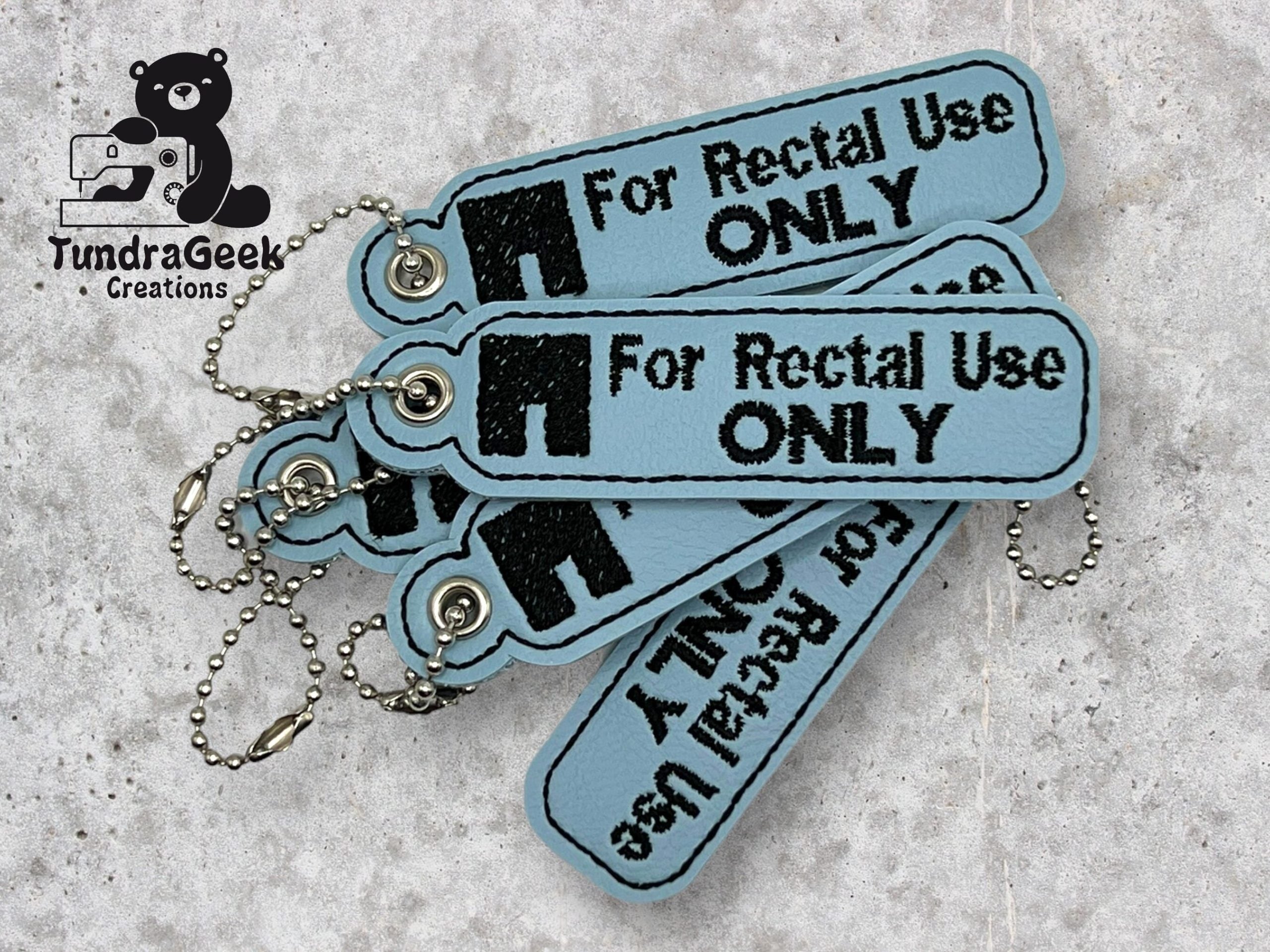 Four blue keychains with metal chains display the text For Rectal Use ONLY in black, with a logo reading TundraGeek Creations in the top left corner.