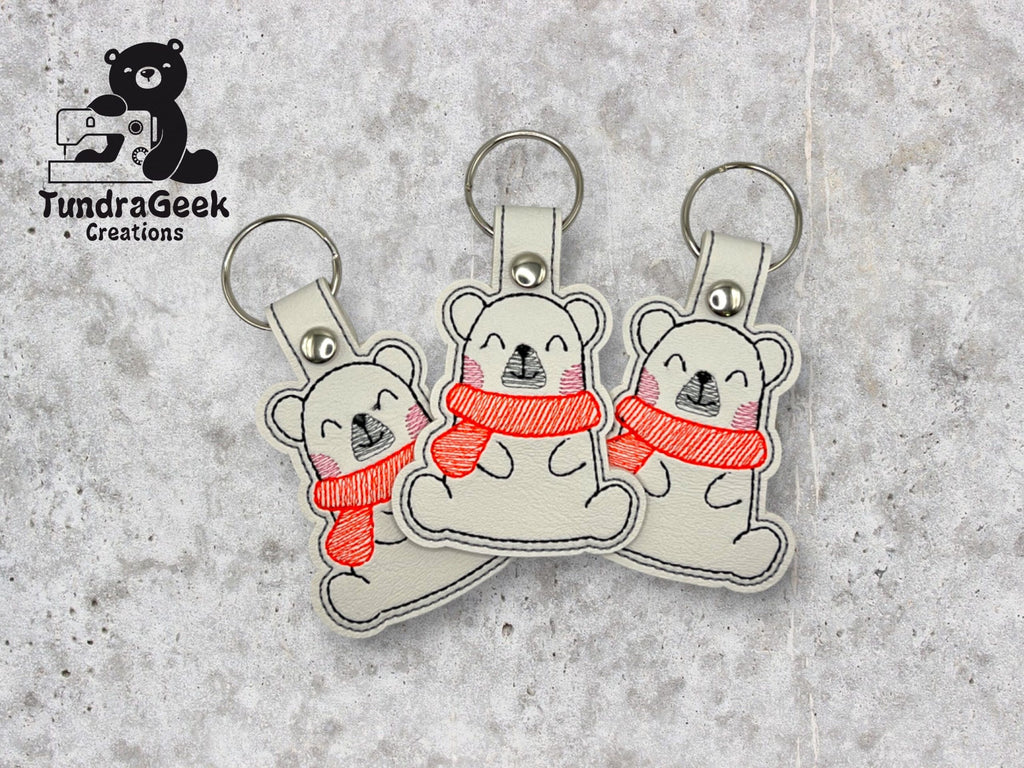 Three gray bear keychains with red scarves, made of stitched fabric, displayed on a stone background with the TundraGeek Creations logo in the top left corner.