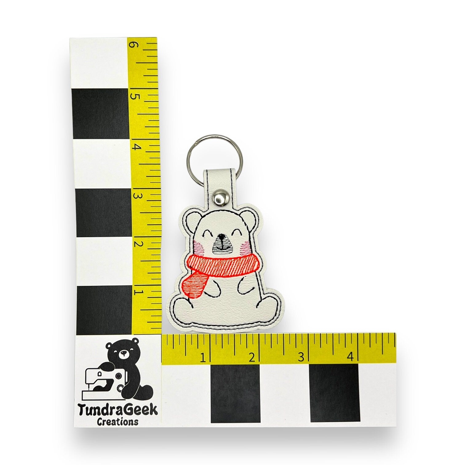 A keychain featuring a bear with a red scarf is positioned against a yellow and black measurement scale labeled “TundraGeek Creations.”.