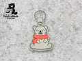A keychain featuring a cartoon bear wearing a red scarf sits on a light textured background. TundraGeek Creations logo appears in the top left corner.
