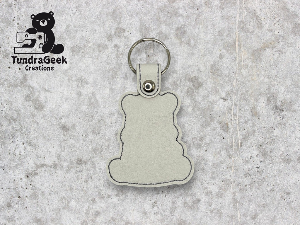 A light gray bear-shaped keychain with black stitching lies on a speckled gray surface. TundraGeek Creations logo is visible in the top left corner.