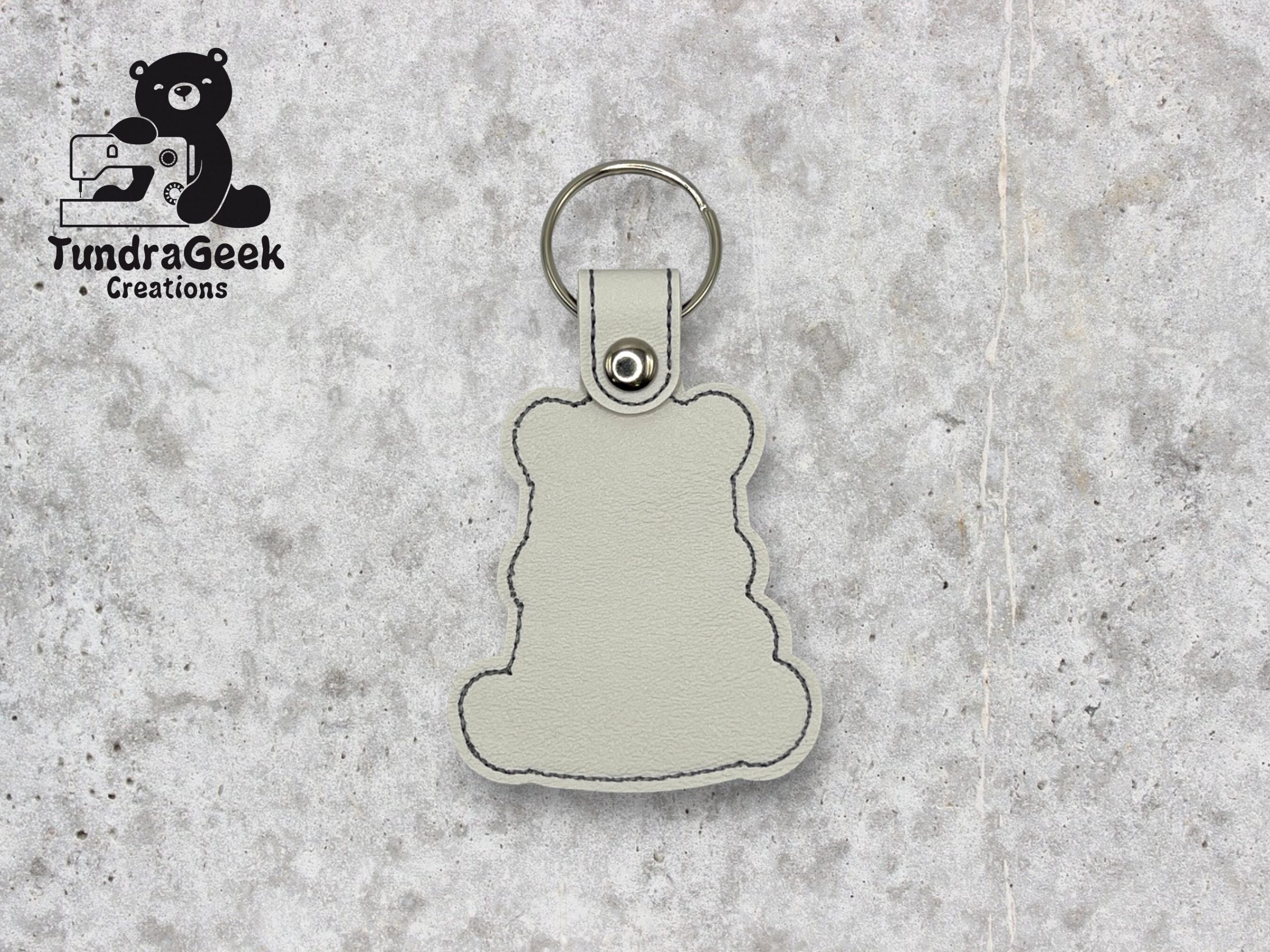 A light gray bear-shaped keychain with black stitching lies on a speckled gray surface. TundraGeek Creations logo is visible in the top left corner.