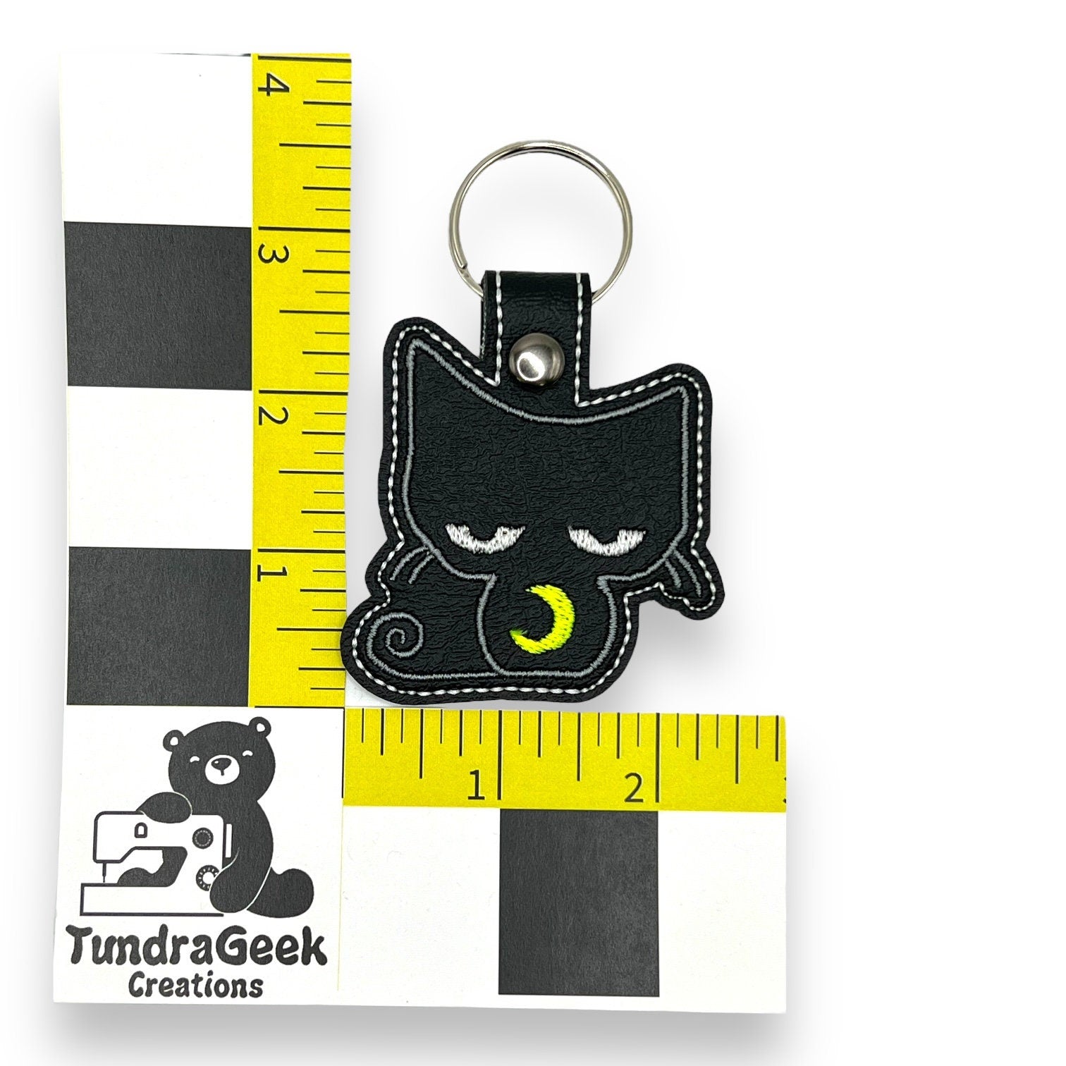 A black cat keychain with closed eyes and a yellow crescent moon, shown next to a ruler for scale, measuring about 3 inches tall.
