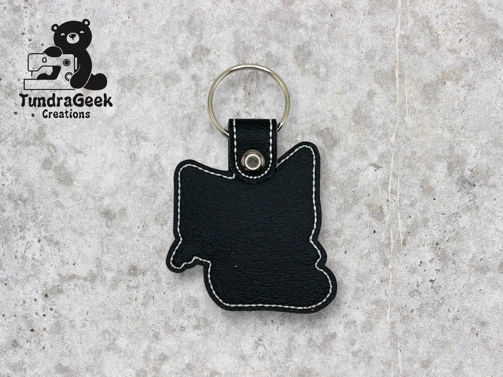 A black leather keychain shaped like the state of Minnesota with white stitching, lying on a gray surface. TundraGeek Creations logo appears in the top left corner.