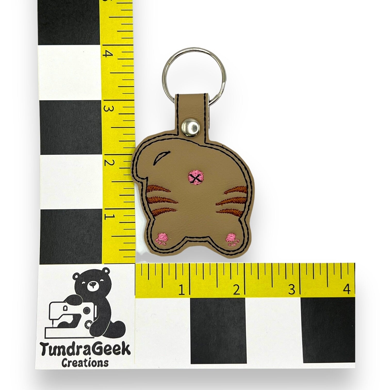 A brown keychain shaped like a cats back, with pink accents, placed next to a ruler showing a size of about 3 inches. TundraGeek Creations logo visible in the corner.