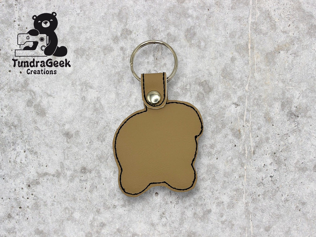 A tan, bear-shaped leather keychain with black stitching, attached to a metal keyring. TundraGeek Creations logo is in the top left corner.
