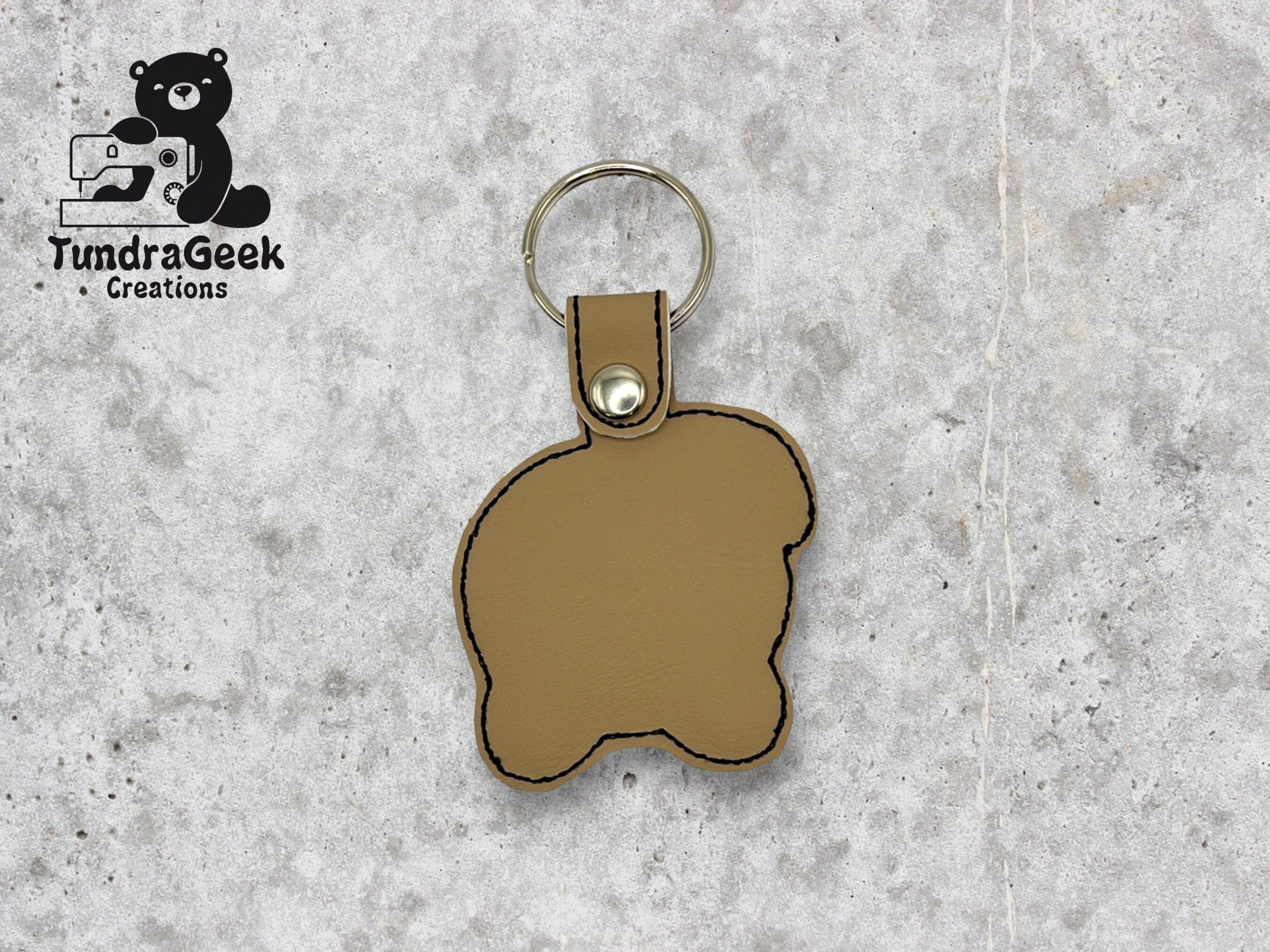 A tan, bear-shaped leather keychain with black stitching, attached to a metal keyring. TundraGeek Creations logo is in the top left corner.