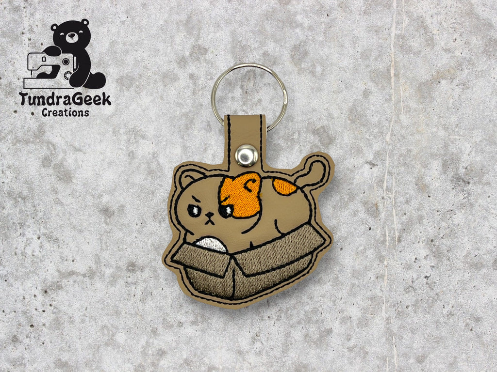 Embroidered keychain featuring a brown cat with an orange patch sitting in a cardboard box, displayed on a light gray textured background. TundraGeek Creations logo in the top left corner.