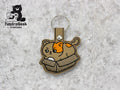 Embroidered keychain featuring a brown cat with an orange patch sitting in a cardboard box, displayed on a light gray textured background. TundraGeek Creations logo in the top left corner.