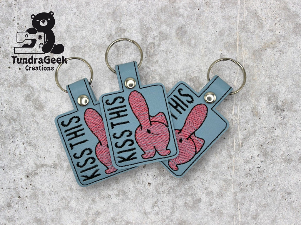Three blue square keychains with embroidered pink cartoon bunnies and the text KISS THIS are displayed on a grey textured surface. TundraGeek Creations logo is in the top left corner.