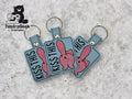 Three blue square keychains with embroidered pink cartoon bunnies and the text KISS THIS are displayed on a grey textured surface. TundraGeek Creations logo is in the top left corner.