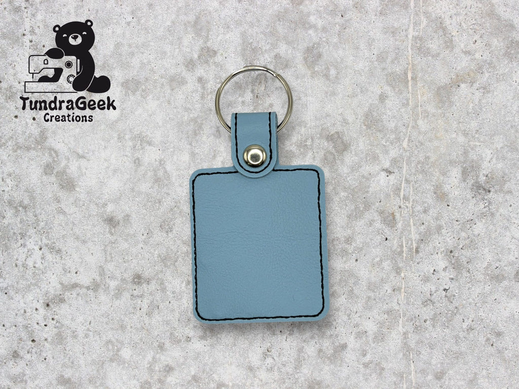 A light blue rectangular leather keychain with black stitching and a metal ring, displayed on a gray textured surface. TundraGeek Creations logo is in the top left corner.
