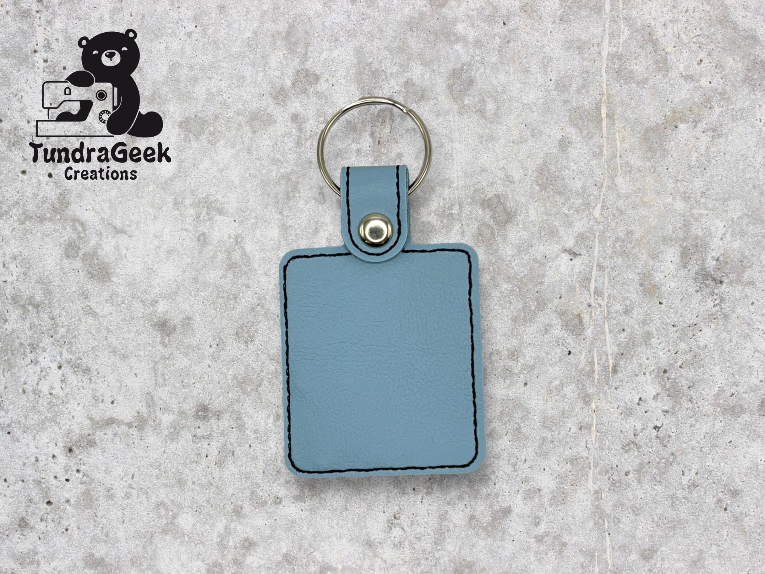 A light blue rectangular leather keychain with black stitching and a metal ring, displayed on a gray textured surface. TundraGeek Creations logo is in the top left corner.