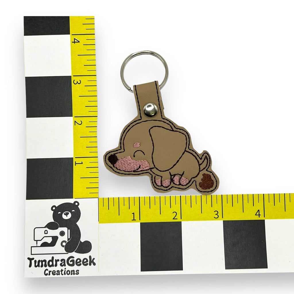 A keychain featuring an embroidered cartoon brown dog is placed next to rulers for scale, showing it measures about 3 inches. A “TundraGeek Creations” logo is visible on the ruler.