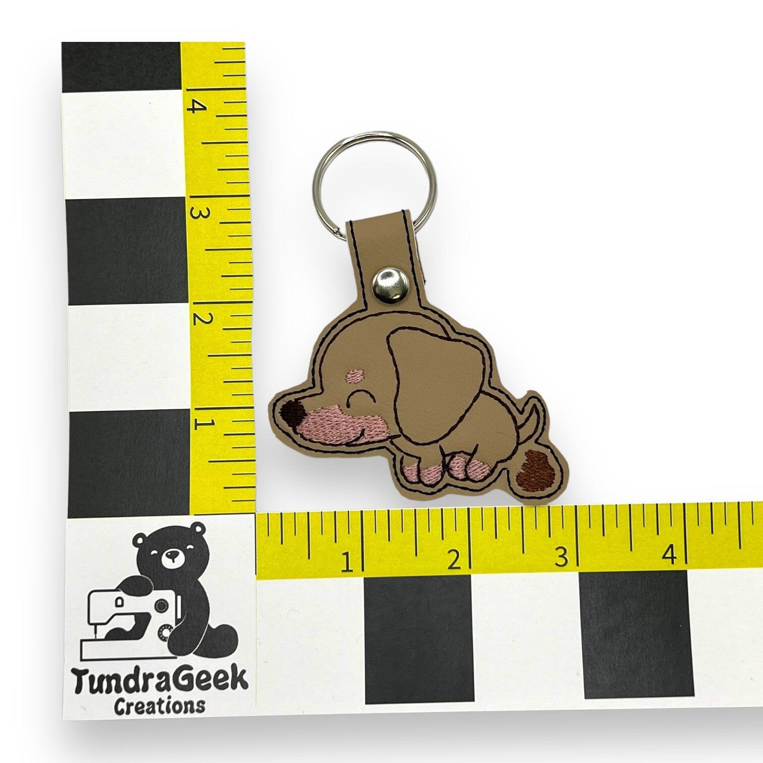 A keychain featuring an embroidered cartoon brown dog is placed next to rulers for scale, showing it measures about 3 inches. A “TundraGeek Creations” logo is visible on the ruler.