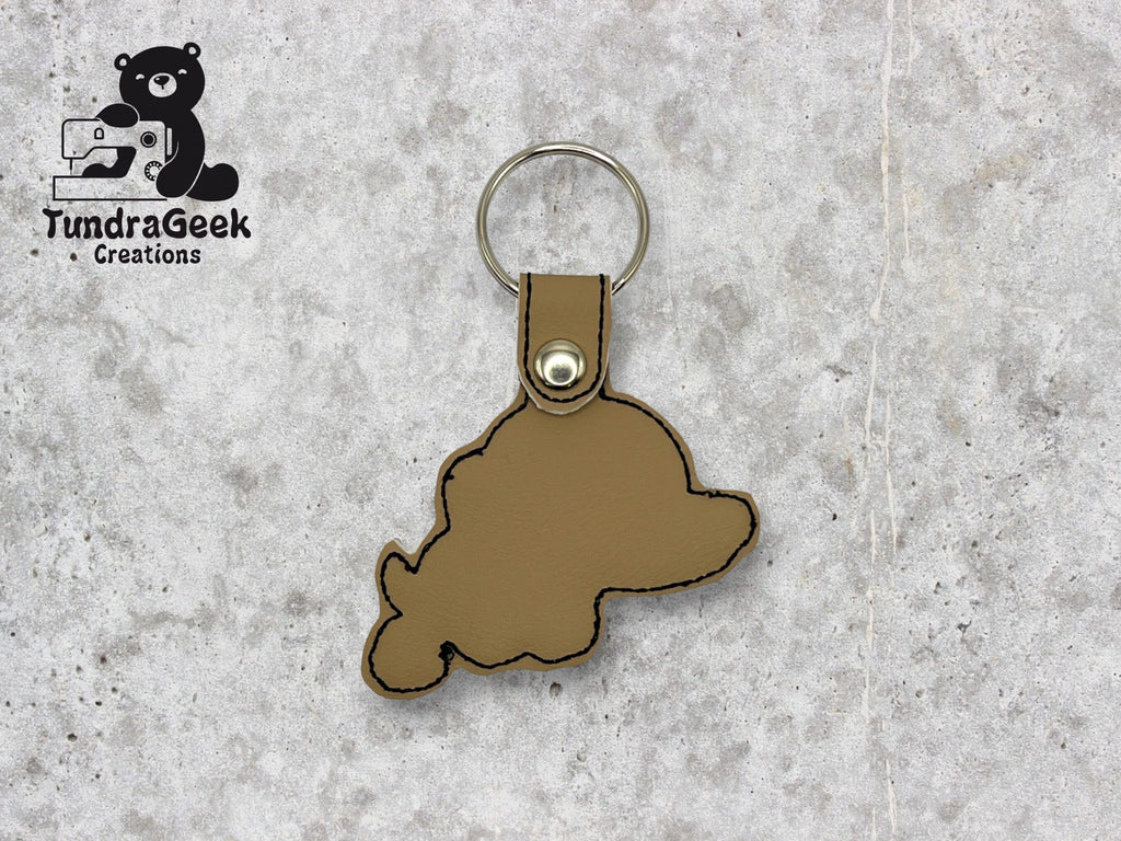 A brown leather keychain in the shape of Nunavut, Canada, with black stitching and a metal keyring, displayed on a gray textured surface. Logo: TundraGeek Creations.