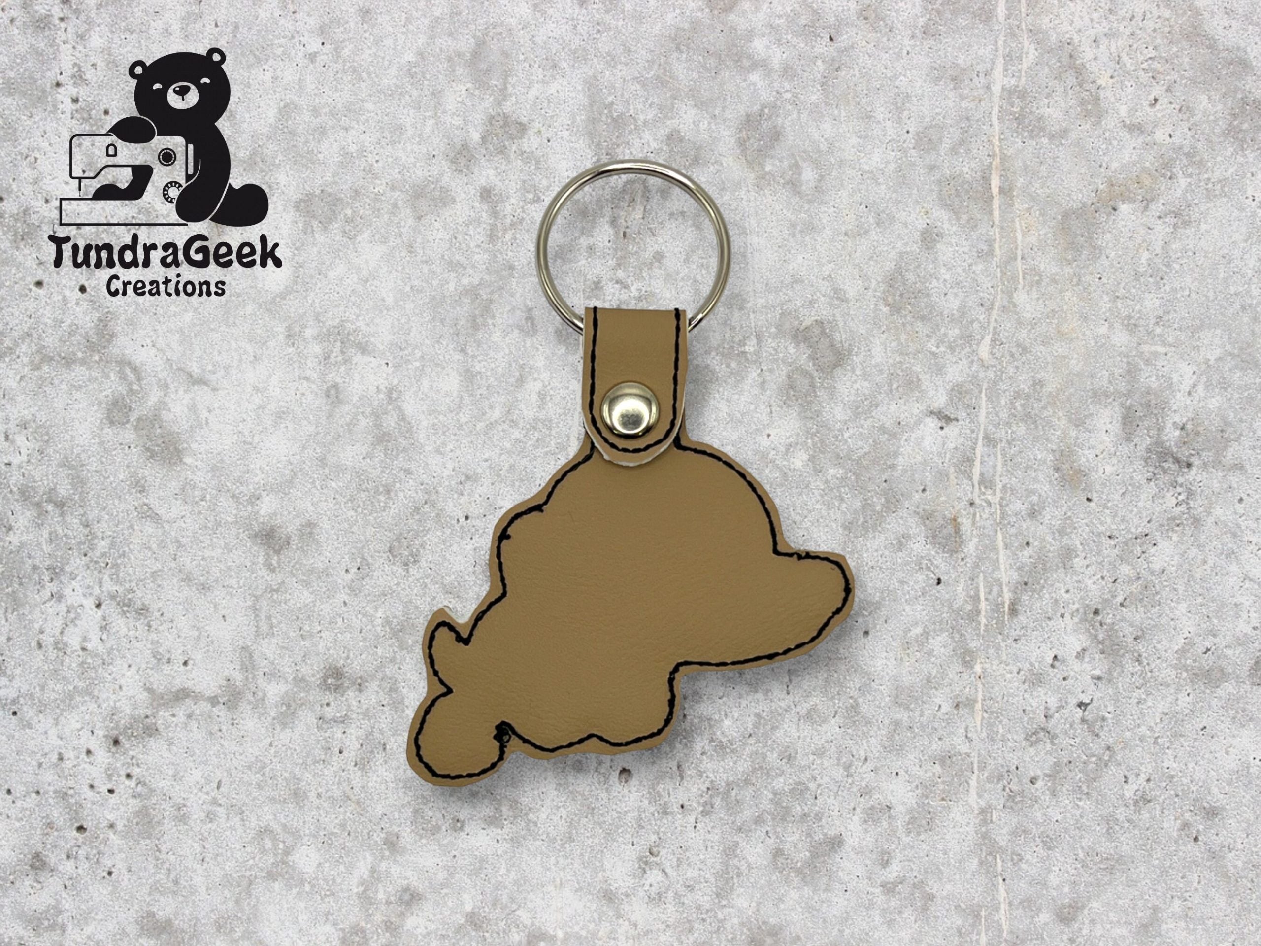 A brown leather keychain in the shape of Nunavut, Canada, with black stitching and a metal keyring, displayed on a gray textured surface. Logo: TundraGeek Creations.