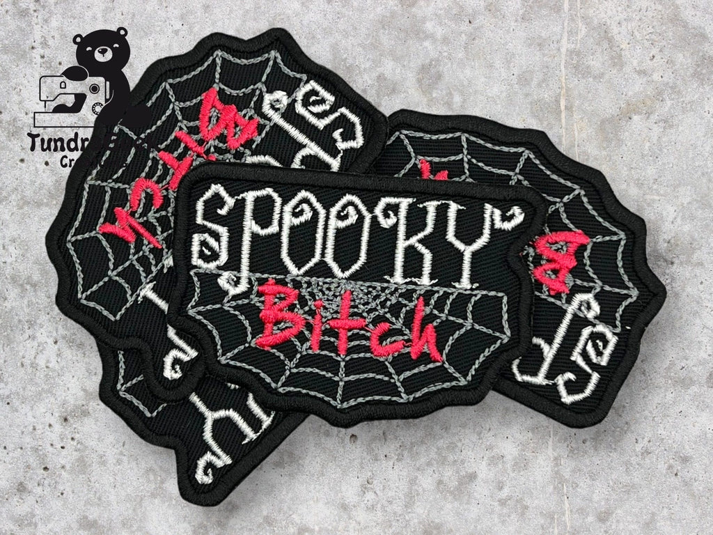 Three black patches with white spiderweb designs and the words SPOOKY Bitch embroidered in white and red thread are arranged on a gray surface.