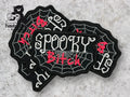 Three black patches with white spiderweb designs and the words SPOOKY Bitch embroidered in white and red thread are arranged on a gray surface.