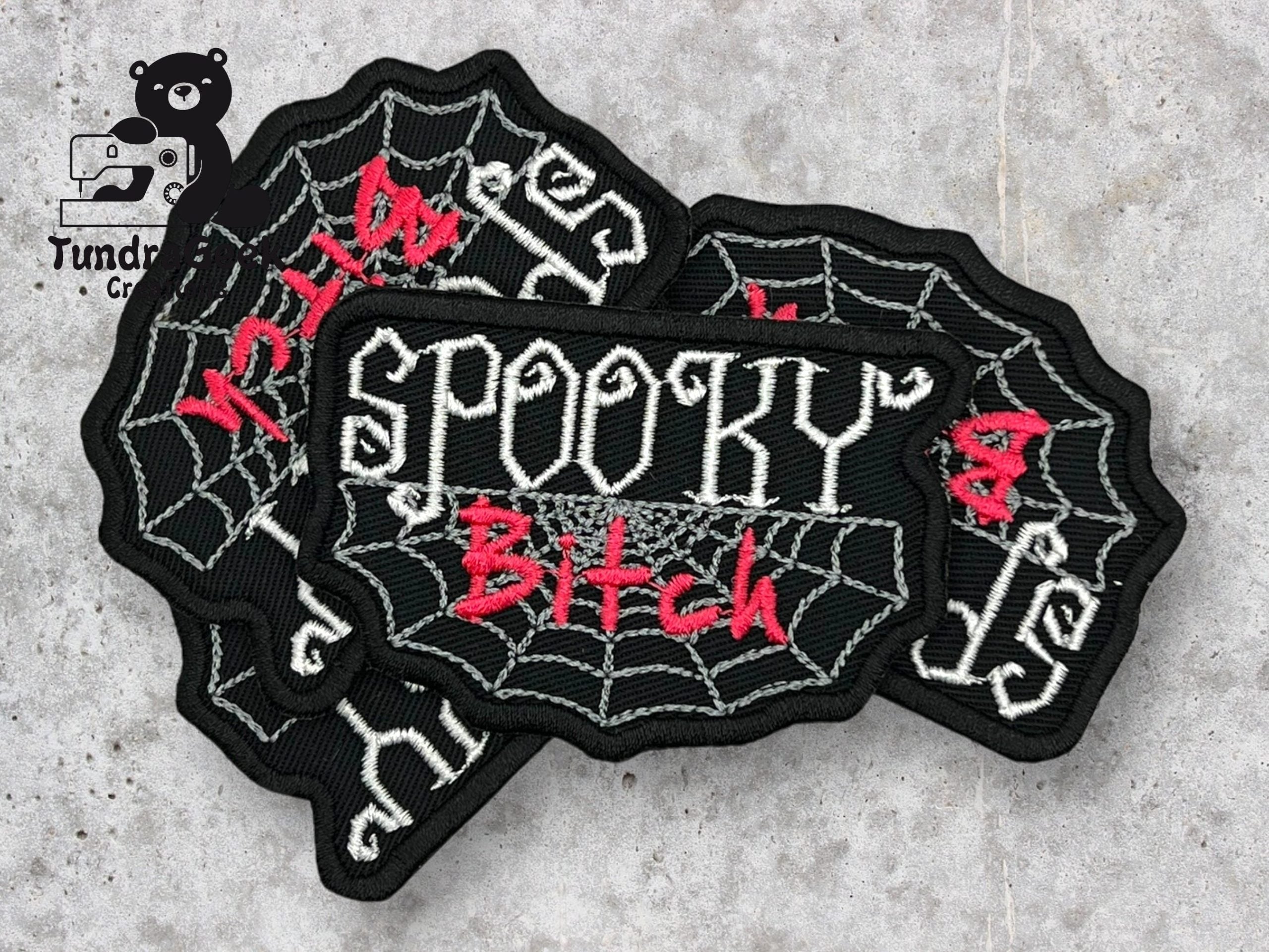 Three black patches with white spiderweb designs and the words SPOOKY Bitch embroidered in white and red thread are arranged on a gray surface.