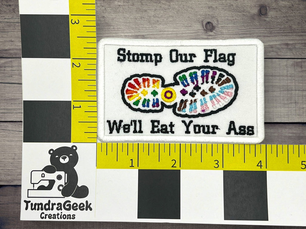 A rectangular embroidered patch with the text Stomp Our Flag Well Eat Your Ass and a colorful footprint; rulers show the patch is about 4 inches wide.
