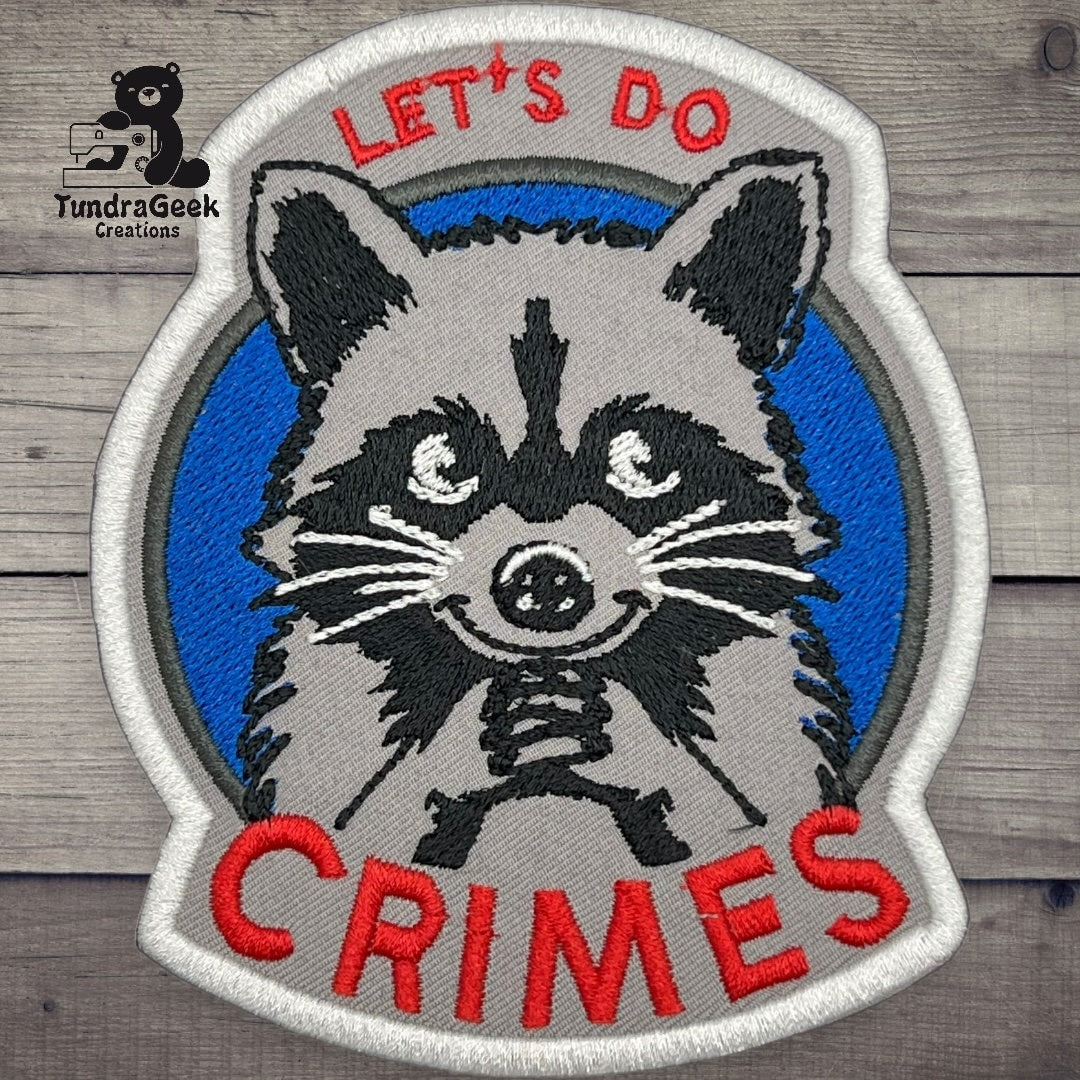 Embroidered patch of a raccoon with clasped paws, blue background, and the words LETS DO CRIMES in red and white letters.