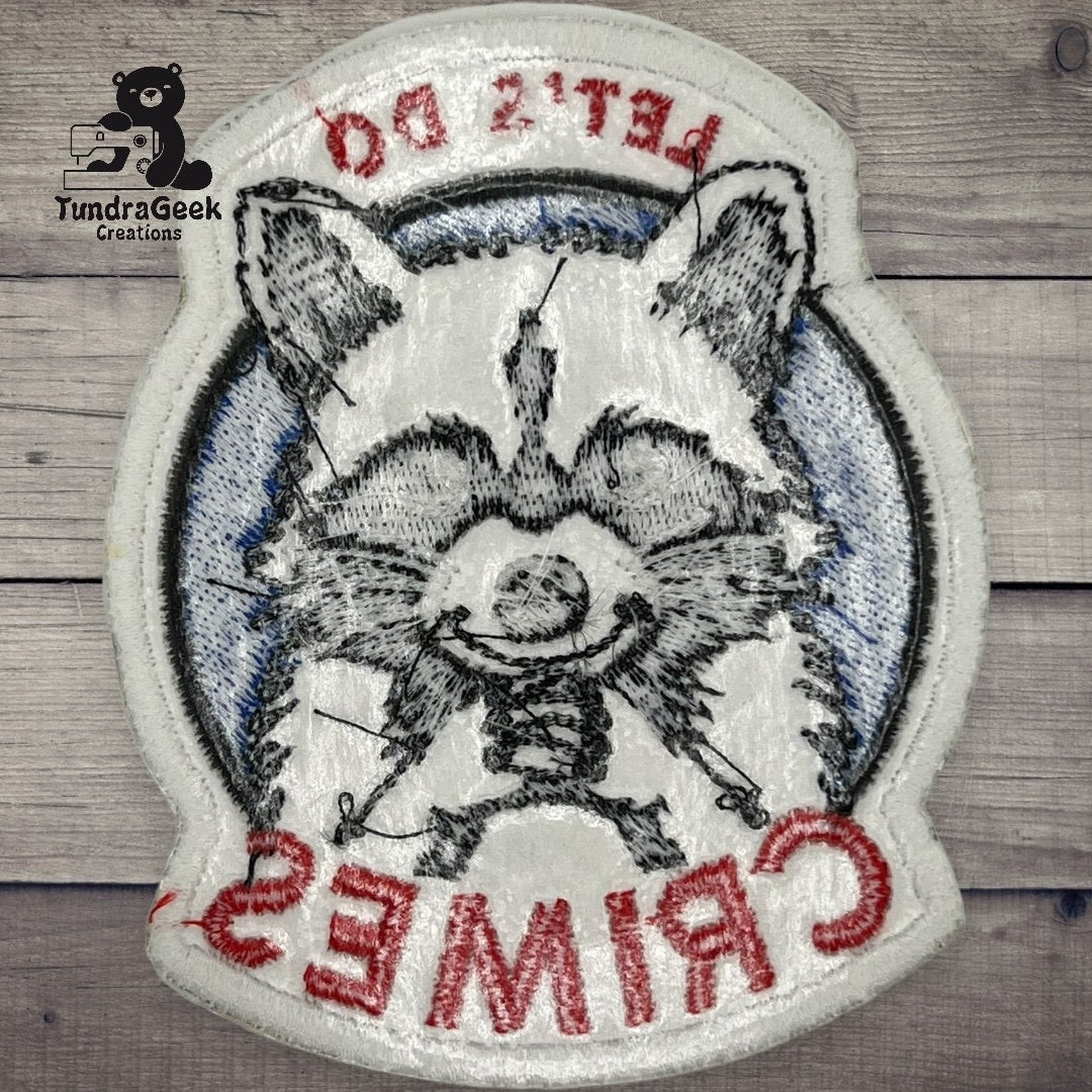 Embroidered patch featuring a raccoon wearing a mask, with the words LETS DO CRIMES and the TundraGeek Creations logo in the corner, on a wooden background.