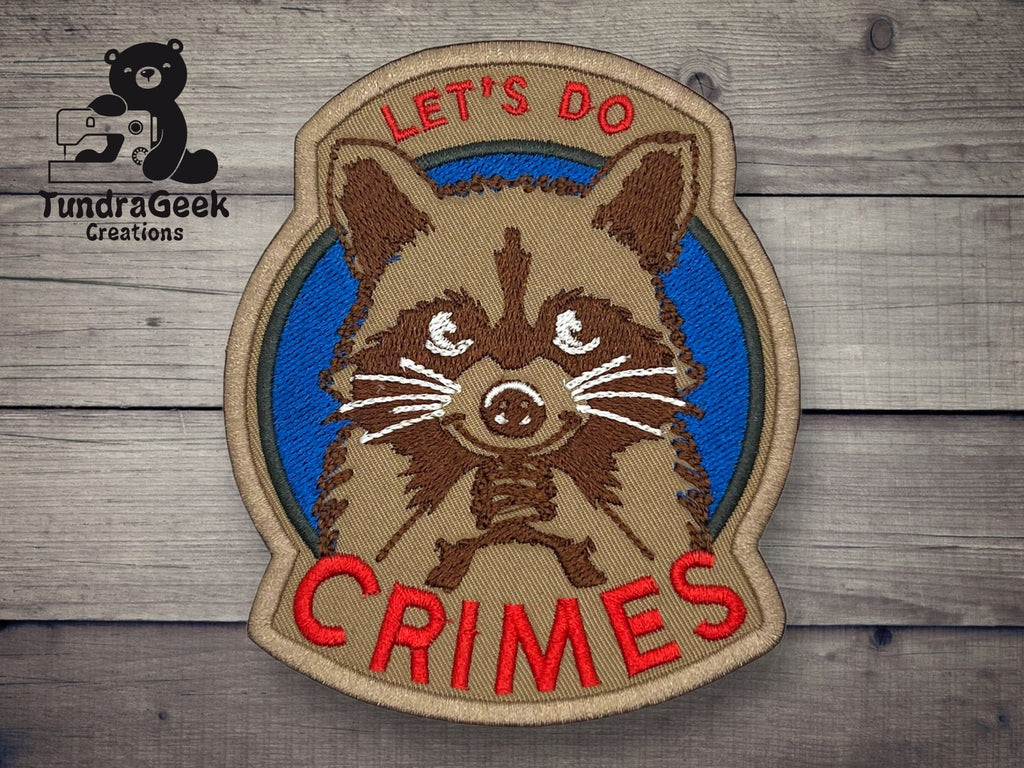 An embroidered patch shows a raccoon with the text LETS DO CRIMES in red and tan letters on a blue background. The TundraGeek Creations logo is in the corner.